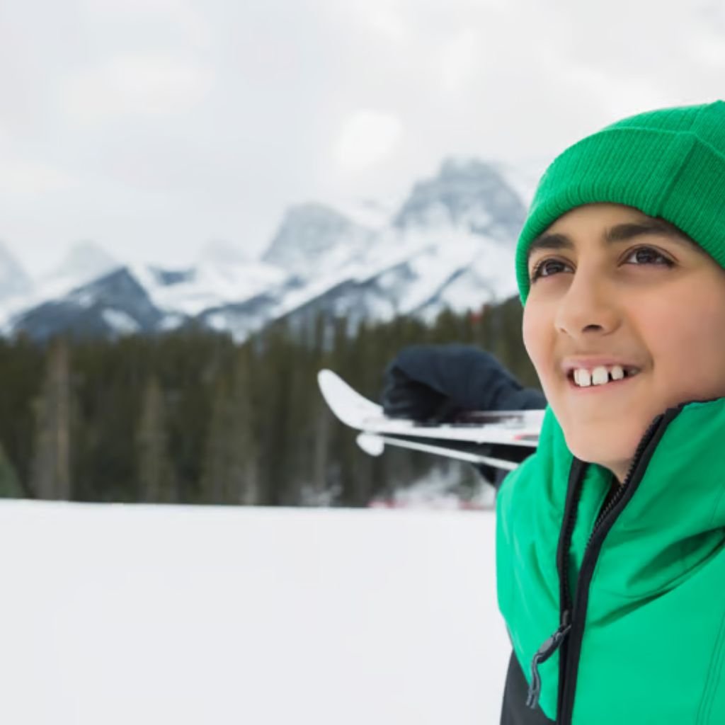 Alt text: Close-up of a young boy wearing a bright green knit hat and a green-and-black winter jacket, smiling as he looks upward. He holds skis over his shoulder, with a snowy field, evergreen forest, and snow-capped mountains in the blurred Snow-Resistant Fabric Explained:background.

