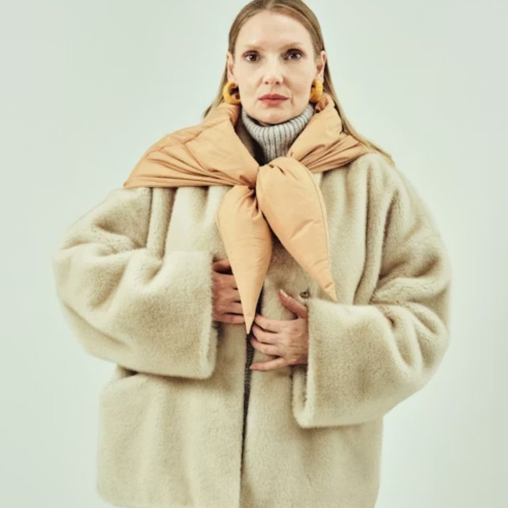 Alt text: A woman wearing an oversized cream-colored fur coat with a peach-toned scarf tied around her shoulders. She has a gray turtleneck underneath and small orange-brown earrings, standing with her hands resting near the coat.
Recycled Fabric Winter Coat:
