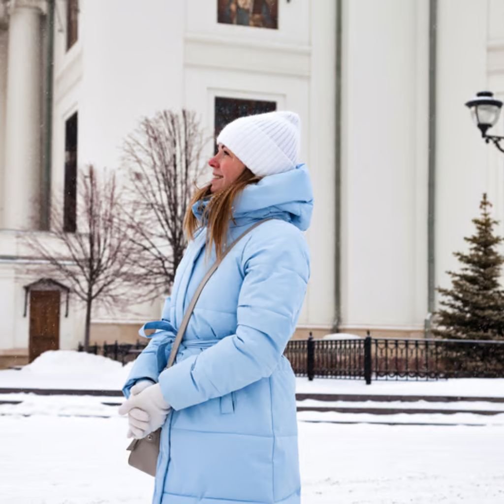 Alt text: A woman standing outdoors in a snowy urban setting, wearing a light blue puffer coat, a white knit beanie, and white gloves. She is looking to the side with a small crossbody bag.

Maximum Warmth Winter Coat Explained: