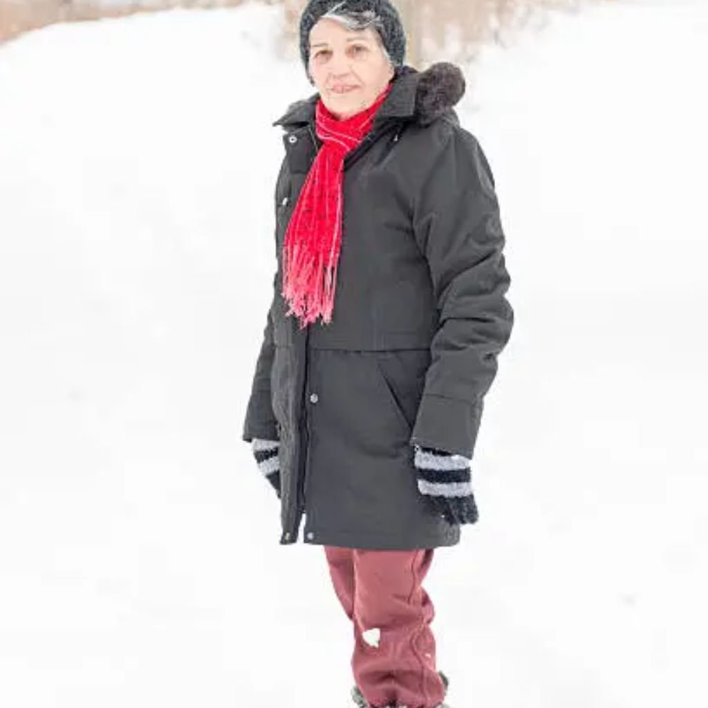 Alt text (short description): A woman standing in a snowy landscape wearing a dark winter coat, red scarf, black hat, striped mittens, and red pants.

Weather-Resistant Winter Coat:
