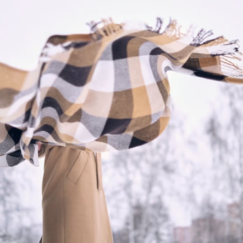 Alt text: Close-up of a person outdoors wearing a large checkered scarf in beige, brown, cream, and gray tones, with a tan coat visible beneath.
Winter Lock Fabric: