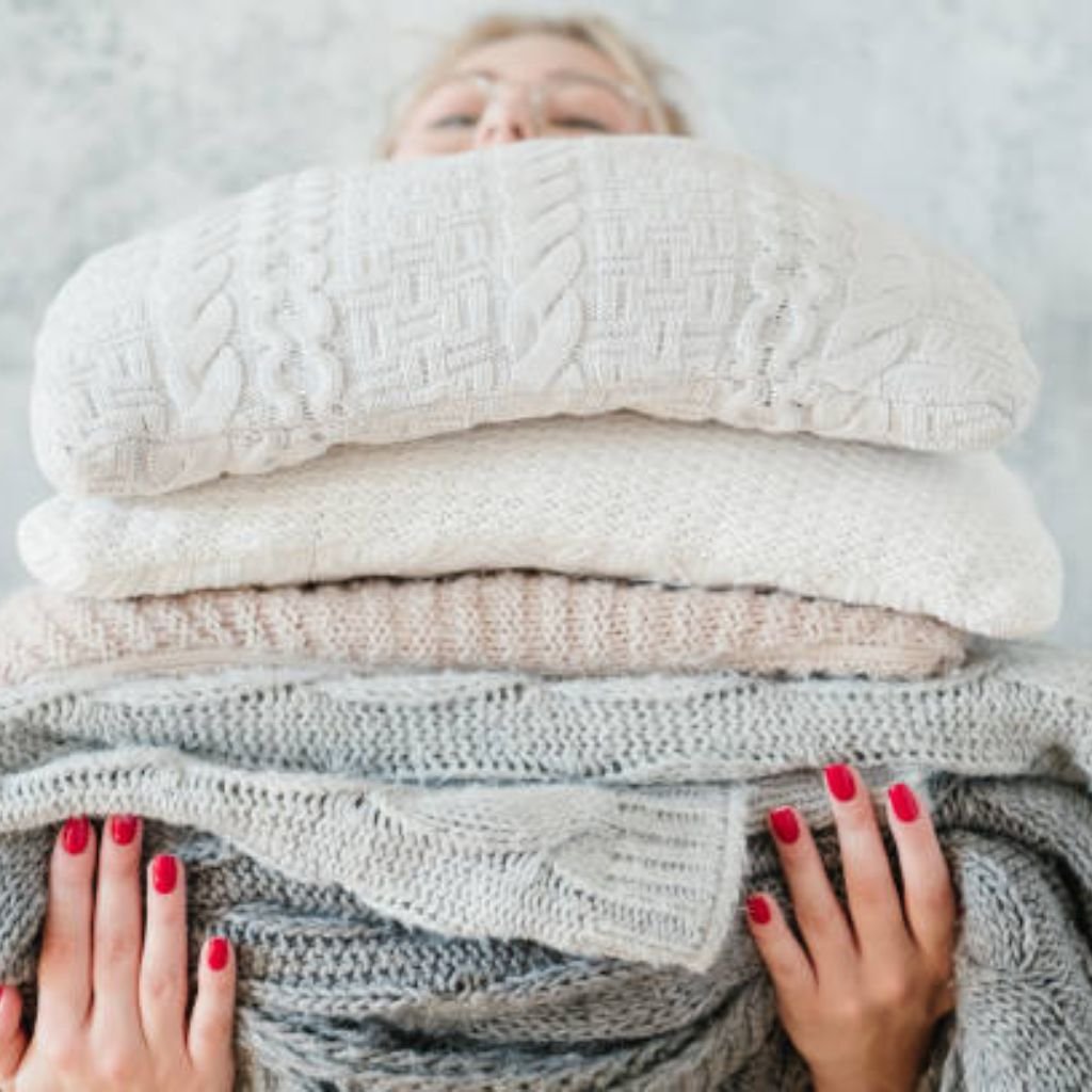 Alt text: A person is partially visible behind a tall stack of folded sweaters in white, cream, light pink, gray, and dark gray. The stack shows varying textures. The person’s forehead, nose, and blurry eyeglasses peek above the top, and their hands with red nail polish hold the sides near the bottom. A light gray textured background surrounds them.

Ever Warm Cozy Fabric Guide: