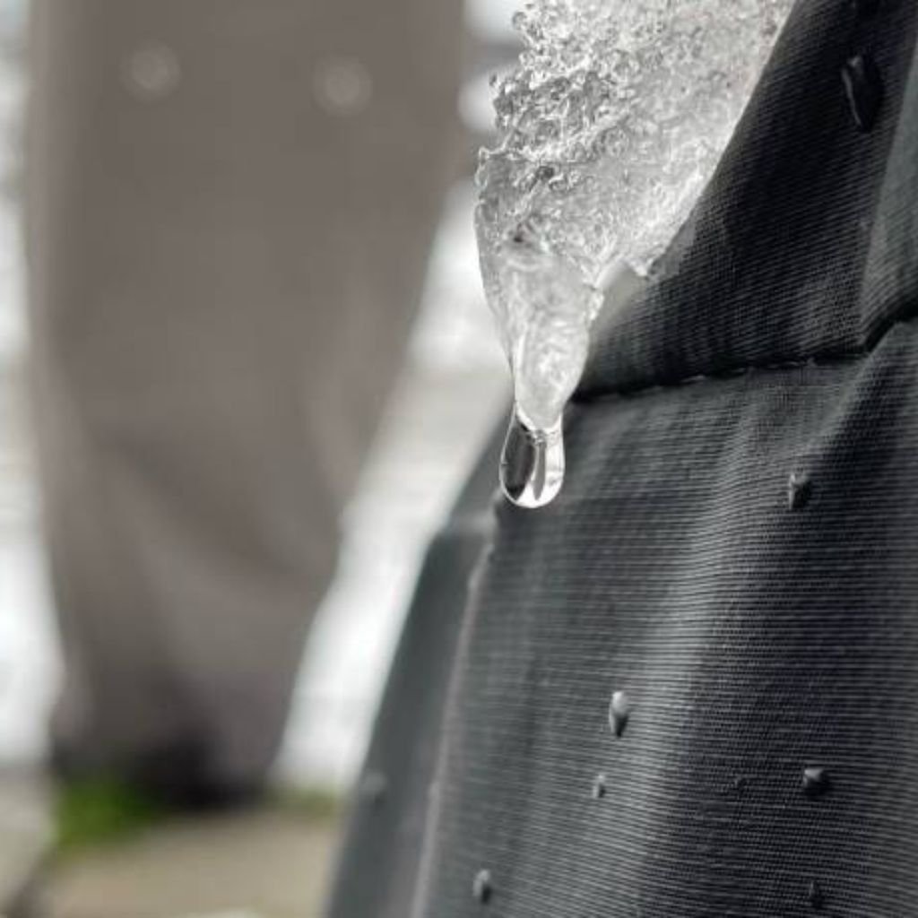 Alt text: Close-up of dark fabric with a melting icicle hanging from its edge and a single water droplet about to fall; a blurred figure stands in the background.

Ice Barrier Windproof Fabric: