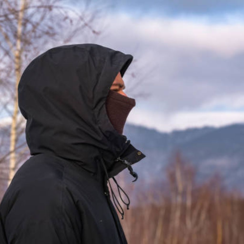 Alt text: A person in a black hooded jacket with a brown face covering visible only around the eyes, standing in a wintery outdoor setting with dry grass and leafless trees, mountains in the distance.

Polar Shield Windproof Knit Explained: