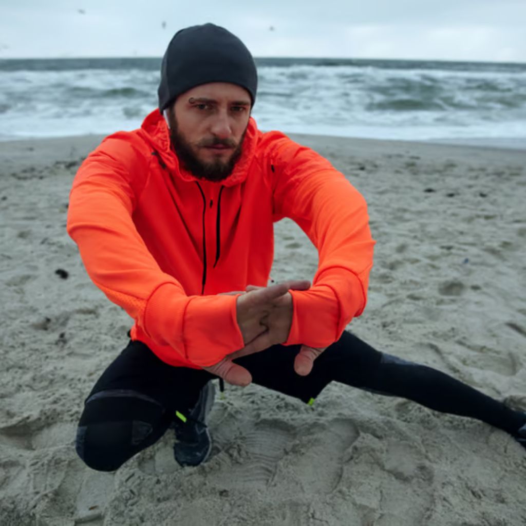 A man in an orange hoodie and black beanie performs a stretch on a sandy beach. The ocean waves are visible in the background as he warms up in athletic gear.
Power Dry Elite Knit Technology: