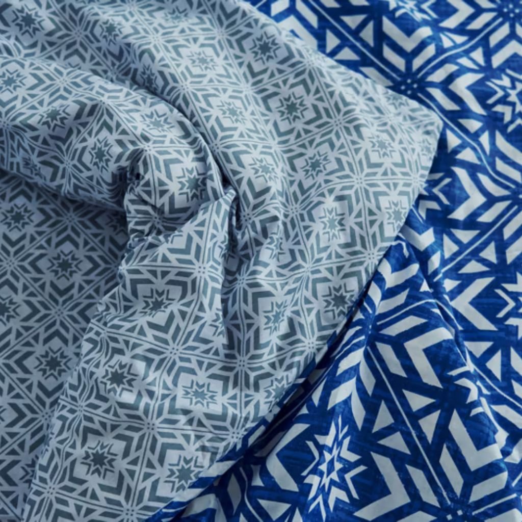 Alt text: Close-up of two folded fabrics in blue tones: a light blue fabric with a star-like geometric pattern on the left and a dark blue fabric with bold white chevron/arrows on the right.
Winter Lock Fabric: