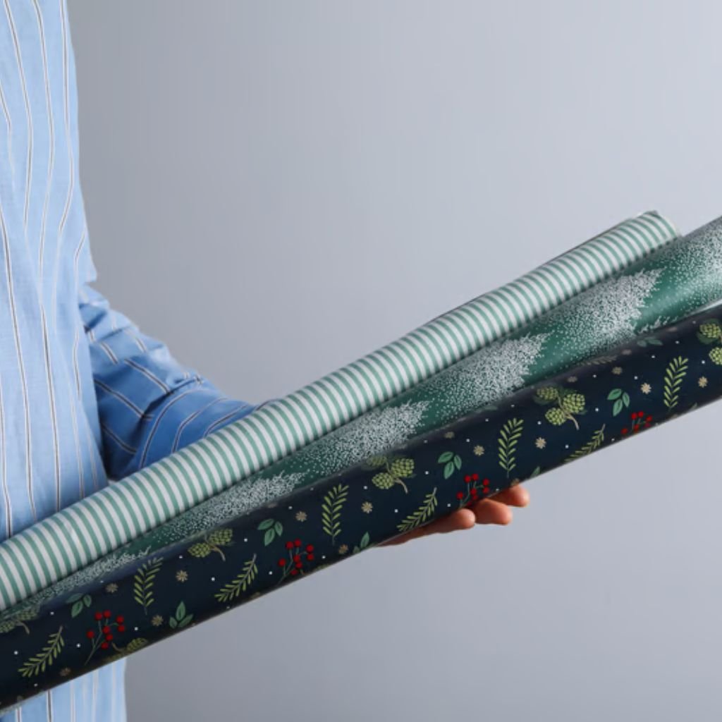Alt text: A person in a blue and white striped shirt holds three rolls of wrapping paper. The top roll is light green with white stripes, the middle roll is dark green with white snowflakes, and the bottom roll is navy blue with red berries, green leaves, and white stars.
Frost Flex Fabric Explained: