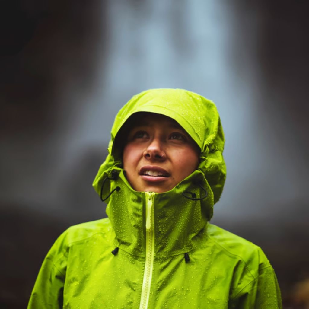 A person wearing a bright green waterproof hooded jacket gazes upward, with droplets of water on the jacket; a blurred natural background suggests rain or mist.

How Gore-Tex Fabric Works: