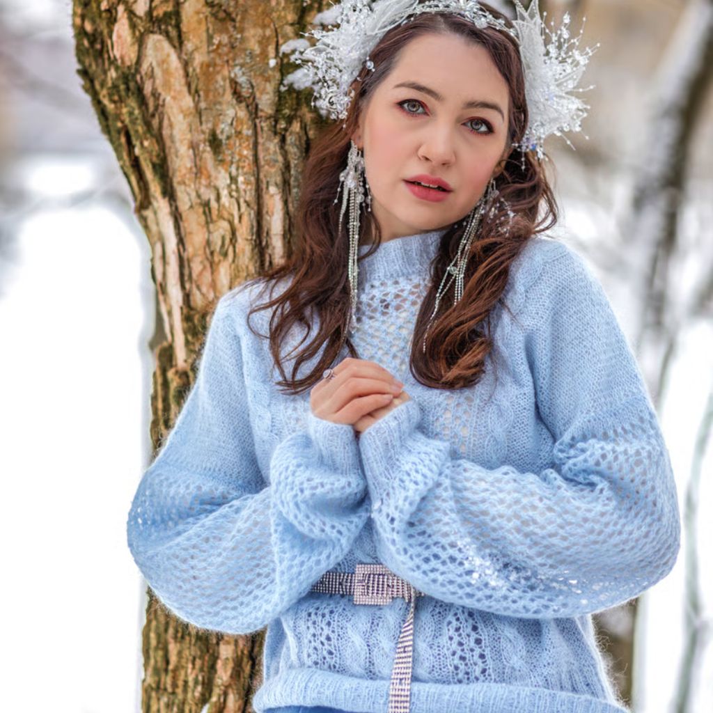 Winter Crown Insul-Fabric Guide: Benefits, Features & Uses 11 A young woman with long brown hair leaning against a tree in a snowy outdoor setting. She wears a light blue open-knit sweater with a silver belt, a decorative silver headpiece, and long sparkling earrings. Her hands are clasped together, and she looks at the camera.Winter Crown Insul-Fabric Guide: