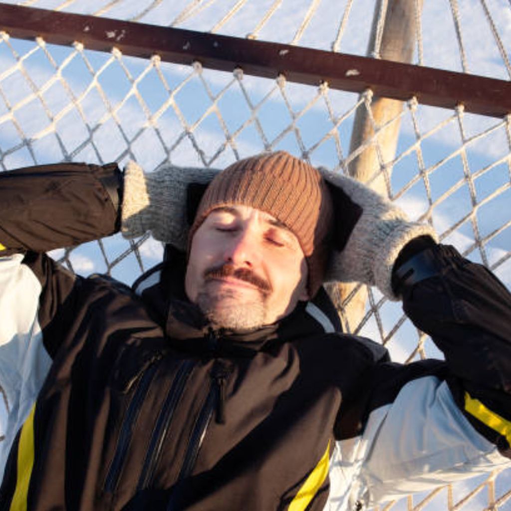 What Is Snow Barrier Climate Fabric? Features & Uses Details 6 Alt text: A man lies in a white net hammock between wooden posts, wearing a brown knit hat, a black jacket with yellow accents, and grey mittens. His eyes are closed and he has a mustache, with a blue sky and clouds in the background.
What Is Snow Barrier Climate Fabric?