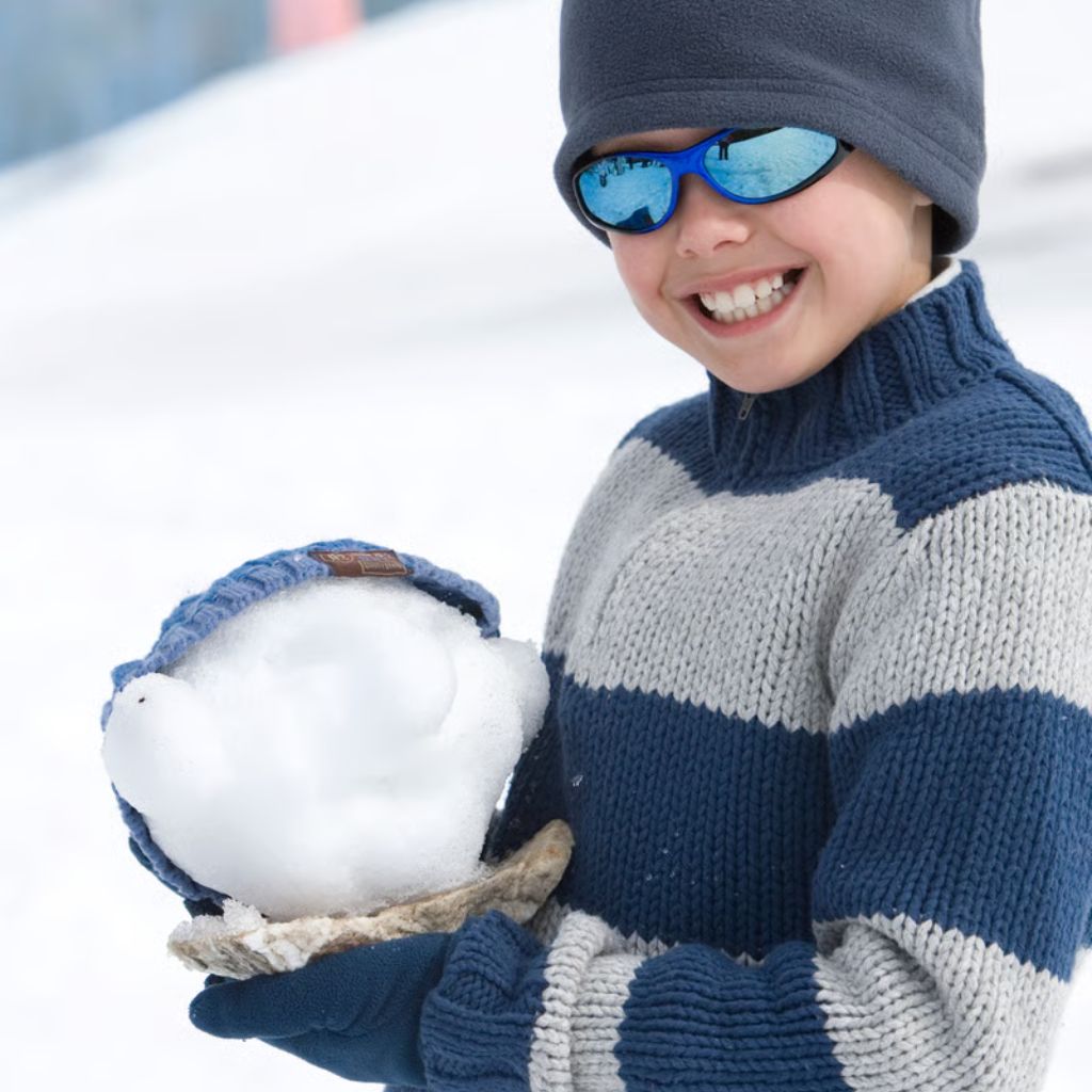 A young boy wearing a blue hat and blue sunglasses holds a large snowball outdoors, wearing a striped blue and gray sweater.
Snow Defense Performance Knit: