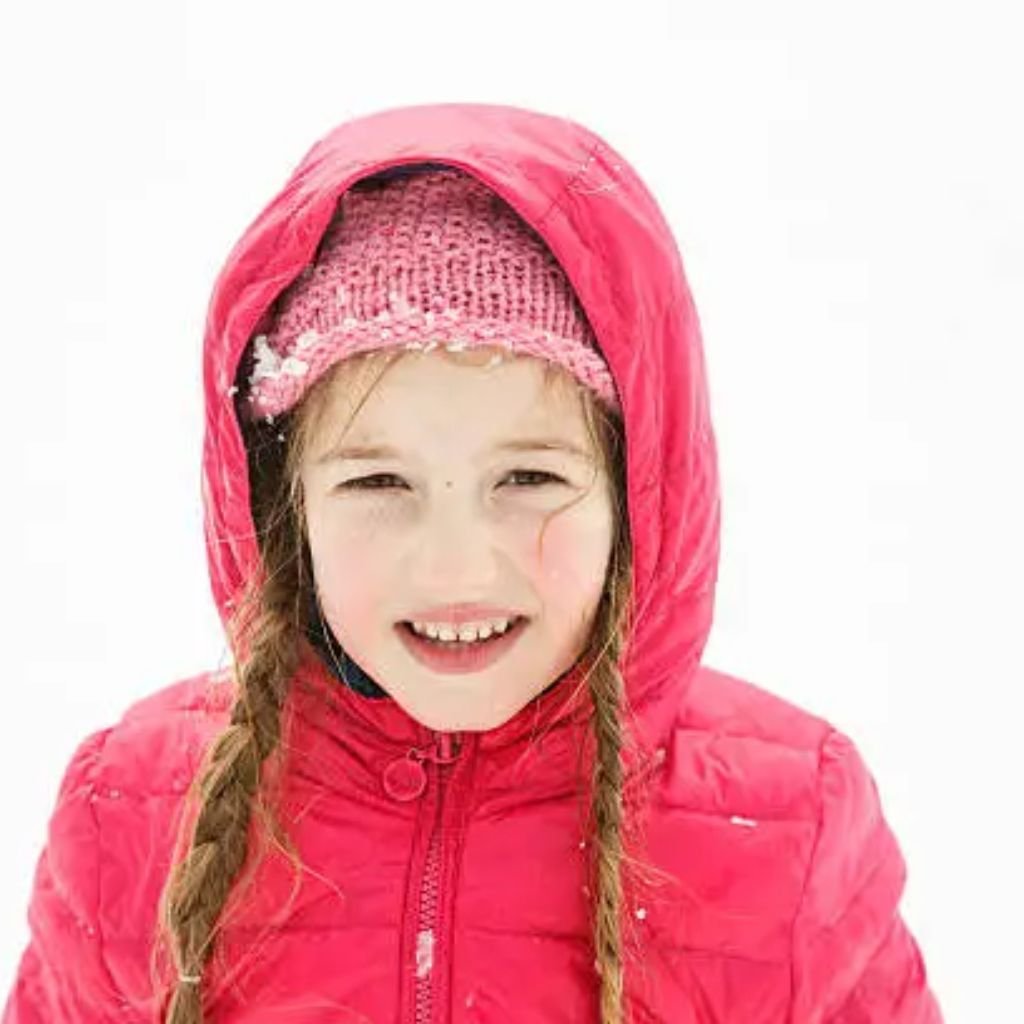 All-Weather Winter Coat: Protection From Rain, Snow & Wind 7 Alt text: A young girl in a bright red hooded winter jacket and a pink knitted hat smiles at the camera. Her braided pigtails peek from under the hood, and there are a few specks of snow on her clothing against a white background.
All-Weather Winter Coat: