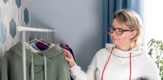 Minimal Logo Hoodie: Sleek, Stylish & Modern Streetwear A woman with blonde hair and glasses stands by a clothing rack, examining a green hoodie among purple and blue garments; she wears a light hoodie with red drawstrings.
