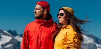 Mountain Rain Jacket Ultimate Protection for Harsh Weather A man and a woman stand together outdoors in a snowy mountainous setting, wearing bright rain jackets (red and yellow) and knit beanies; clear blue sky.