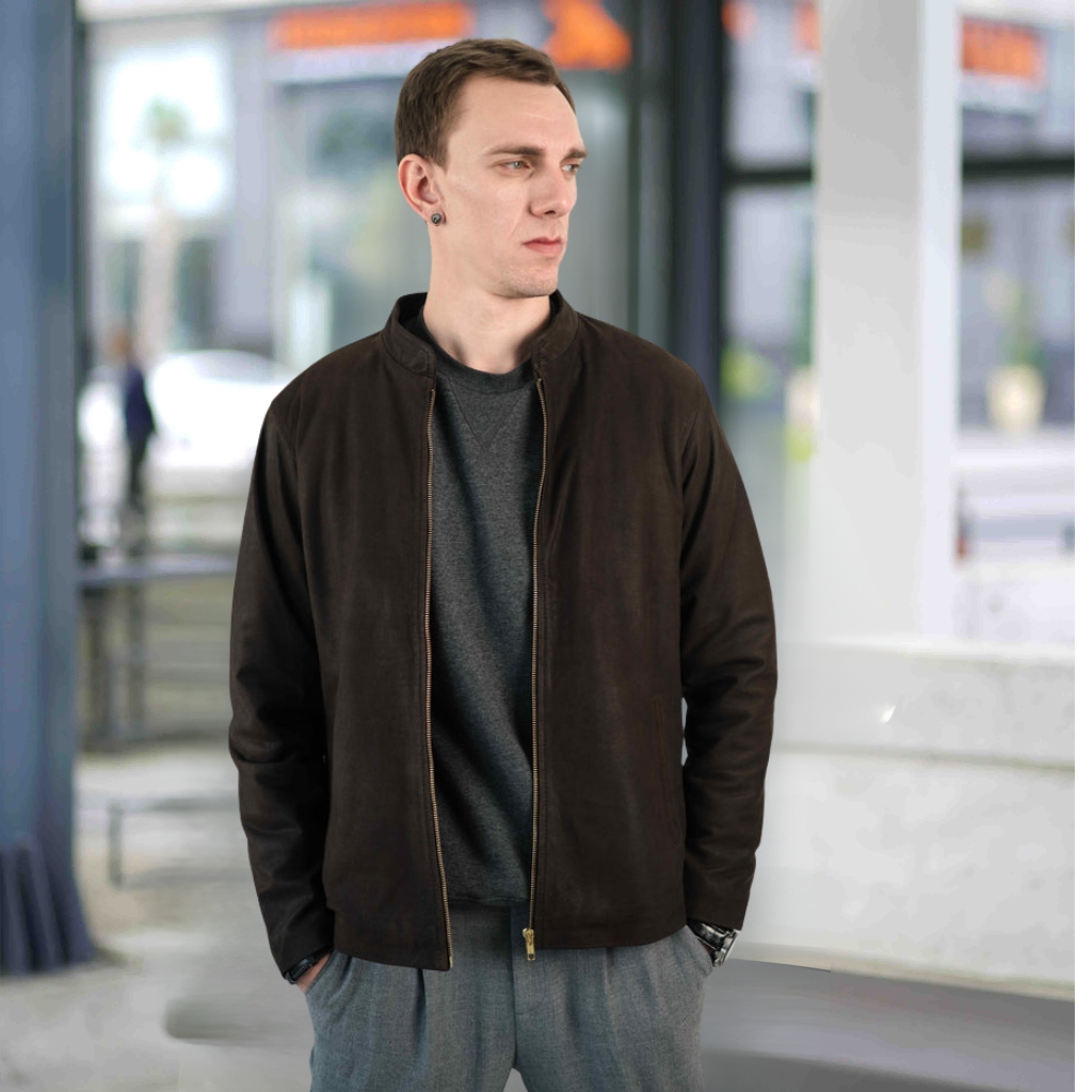 A man stands indoors wearing a dark brown nubuck leather jacket over a gray sweater and gray trousers. He has short brown hair, is looking to his left, and has his hands in his pockets. A watch is visible on his left wrist, and the background shows a blurred interior space with white columns and windows.