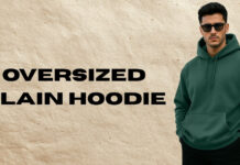 Oversized Plain Hoodie for Effortless Street Style A promotional image featuring a man wearing a dark green oversized plain hoodie with a front kangaroo pocket. He stands on the right, wearing sunglasses and black pants, with his hands in his pockets. The background is a textured light-brown paper-like surface, and bold black text on the left reads “OVERSIZED PLAIN HOODIE.