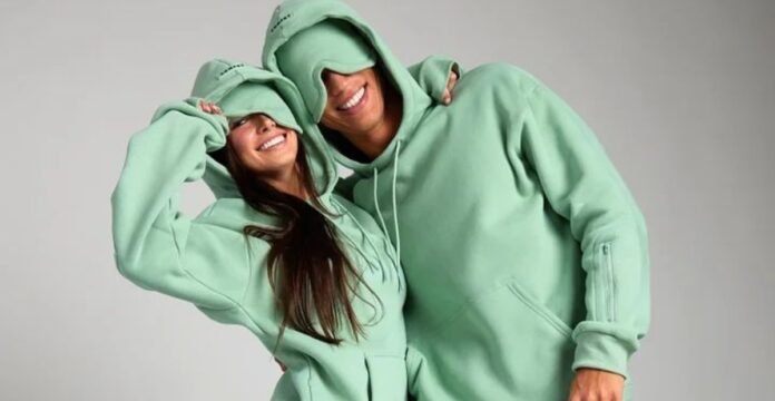 A cheerful photo of a smiling couple wearing matching light-green oversized hoodies. The woman on the left has her hoodie pulled down over her eyes, and the man on the right has his hoodie pulled down as well. They are standing close together against a neutral gray background, embracing playfully.