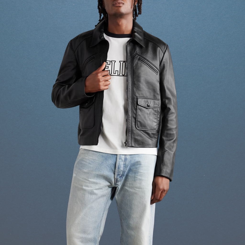 A man stands against a blue backdrop wearing a black leather paneled jacket open over a white graphic T-shirt and light wash jeans. His hair is styled in dreadlocks.