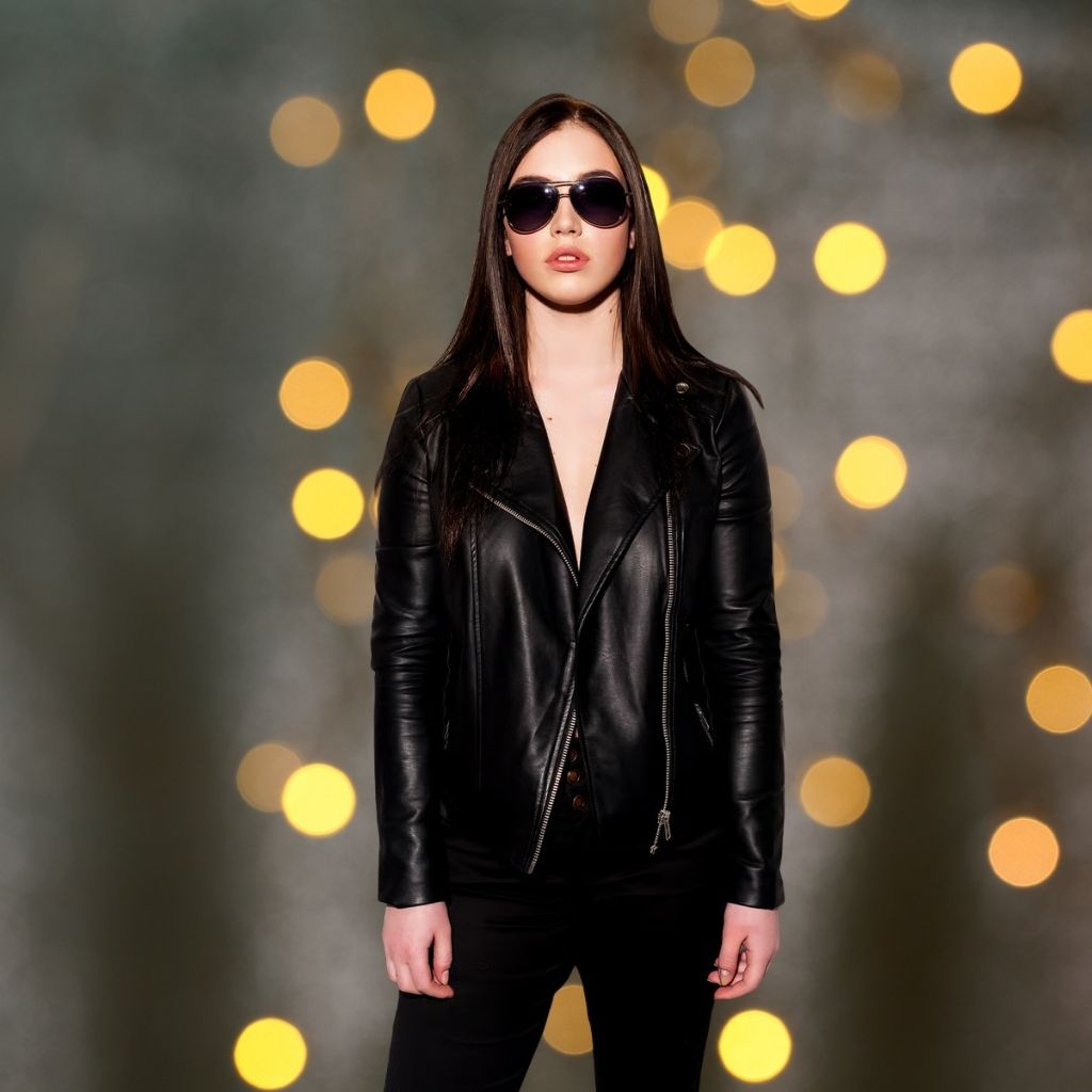 A young woman stands facing the camera in a black leather jacket, black pants, and dark sunglasses. She has long dark hair, and the background features warm golden bokeh lights, giving a festive, evening vibe.Party Leather Jacket