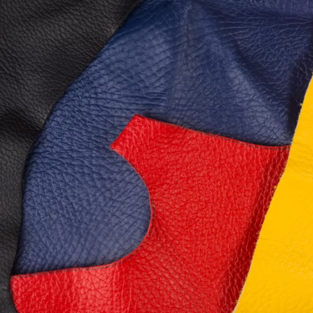 Stylish Patchwork Leather Jacket with Bold Multicolor Design 5 A close-up of an abstract patchwork leather design with overlapping black, blue, red, and yellow patches. The red patch features a curved indentation along its edge where it meets the blue patch, and the yellow patch appears on the right side of the red patch. The leather surfaces show a rich grain texture.
