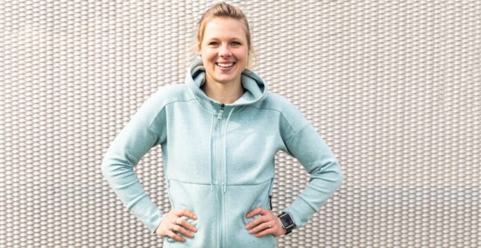 Smiling woman standing with hands on hips in front of a textured white wall, wearing a light blue zip hoodie and a smartwatch on her left wrist.