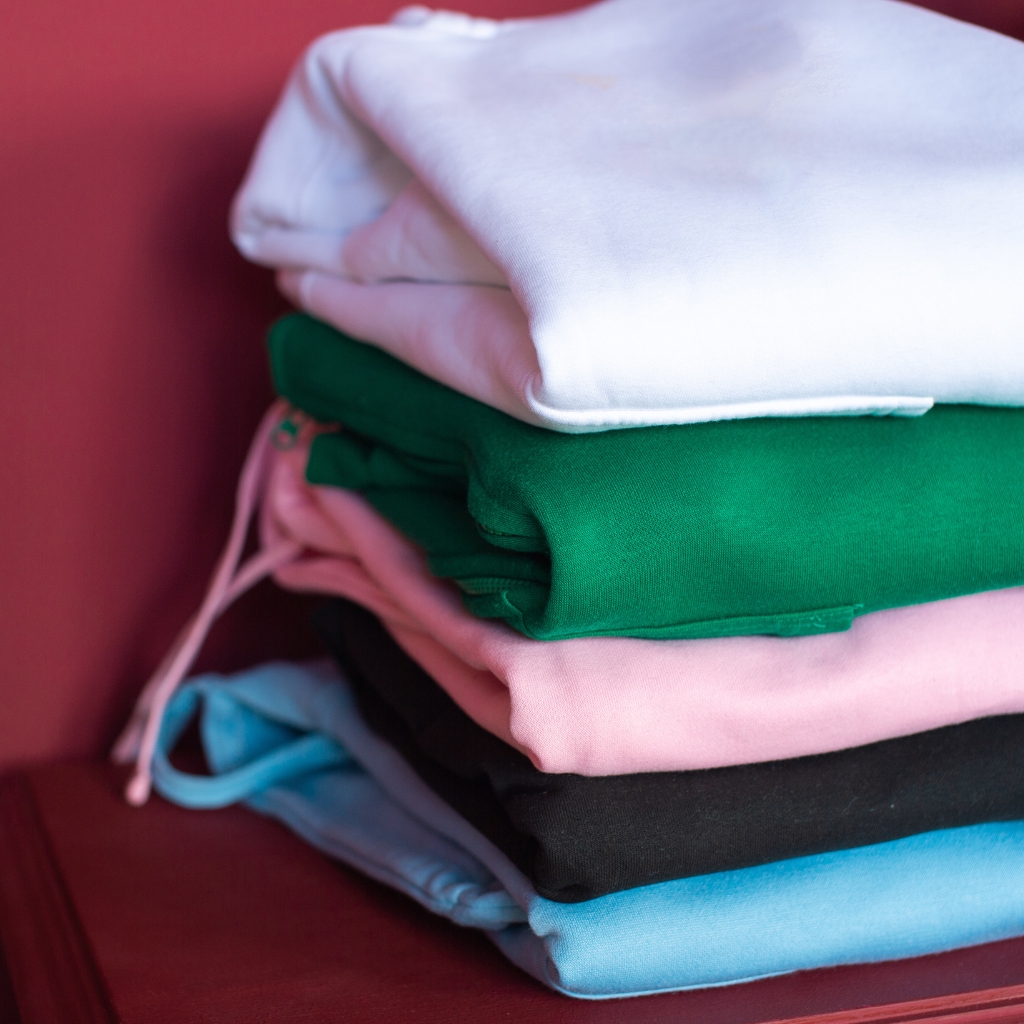 Minimal Logo Hoodie: Sleek, Stylish & Modern Streetwear 3 A stack of folded hooded sweatshirts in white, green, pink, black, and light blue on dark red furniture.