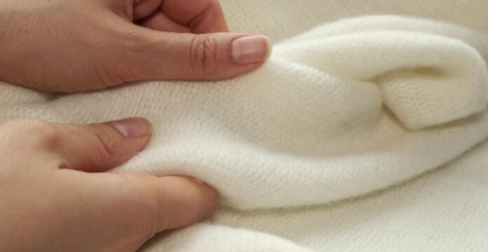 Recycled Cashmere Jersey Fabric Close-up of hands gently handling a soft cream-colored knit fabric with a plush, fuzzy surface.