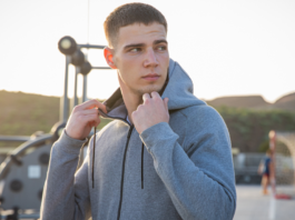 A young man in a grey zip-up hoodie stands outdoors, holding the collar and looking to the side, with an outdoor gym and playground in the background during sunset.