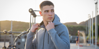 Ripped Zip Hoodie: Bold Streetwear Essential A young man in a grey zip-up hoodie stands outdoors, holding the collar and looking to the side, with an outdoor gym and playground in the background during sunset.