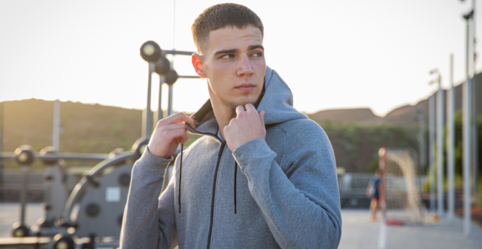 A young man in a grey zip-up hoodie stands outdoors, holding the collar and looking to the side, with an outdoor gym and playground in the background during sunset.