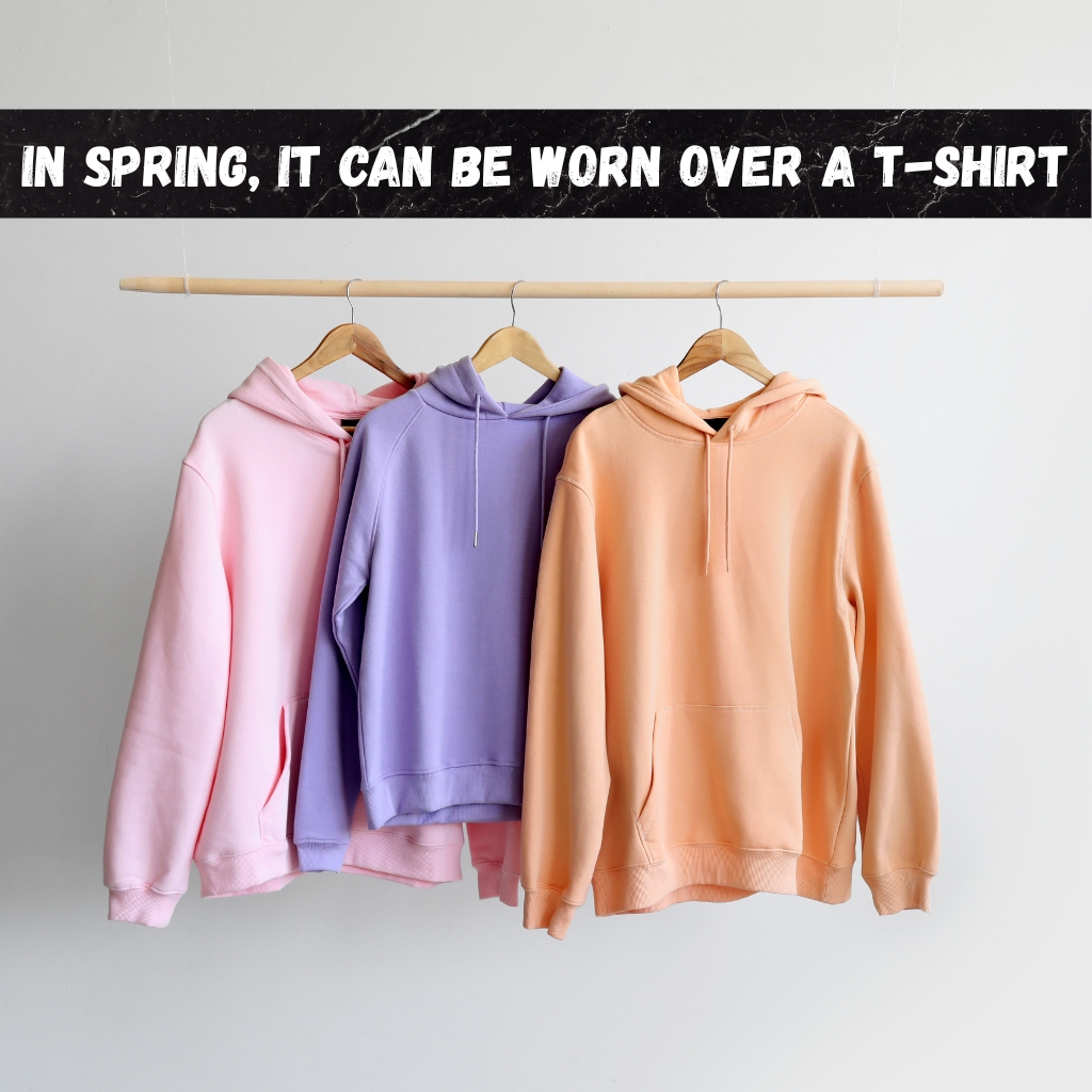 Vintage Zip Hoodie: Classic Retro Streetwear Style 5 Three pastel-colored hooded sweatshirts (pink, lavender, peach) hanging on a wooden rod with wooden hangers against a white background. A black sign with white text above reads 'IN SPRING, IT CAN BE WORN OVER A T-SHIRT.'