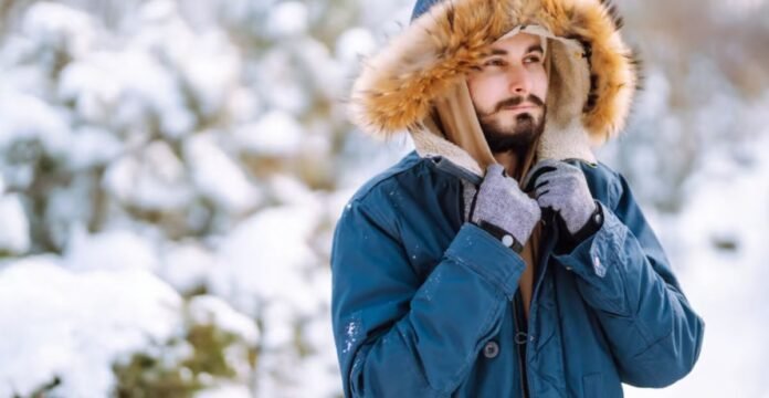 A man outdoors in a snowy setting, wearing a blue parka with a fur-lined hood and gray gloves, adjusting the hood strings. Maximum Warmth Winter Coat: