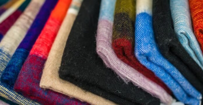 Alt text: Close-up of a neatly stacked pile of folded fabrics in a diagonal arrangement. Colors include blue, red, orange, beige, black, purple, and green with some patterns; the texture appears soft and warm. Sustainable Wool Fabric: