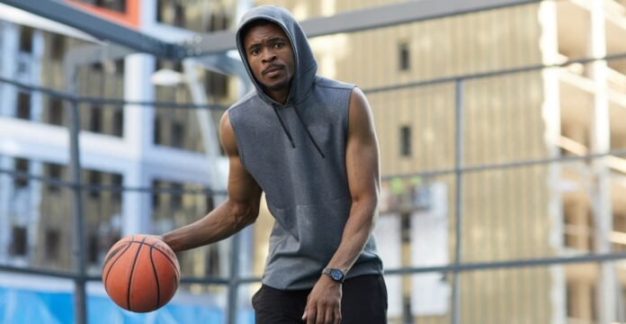 Athletic African-American man in a gray sleeveless hoodie dribbling a basketball on an outdoor court, wearing a watch. Buildings and a wire fence are in the background.