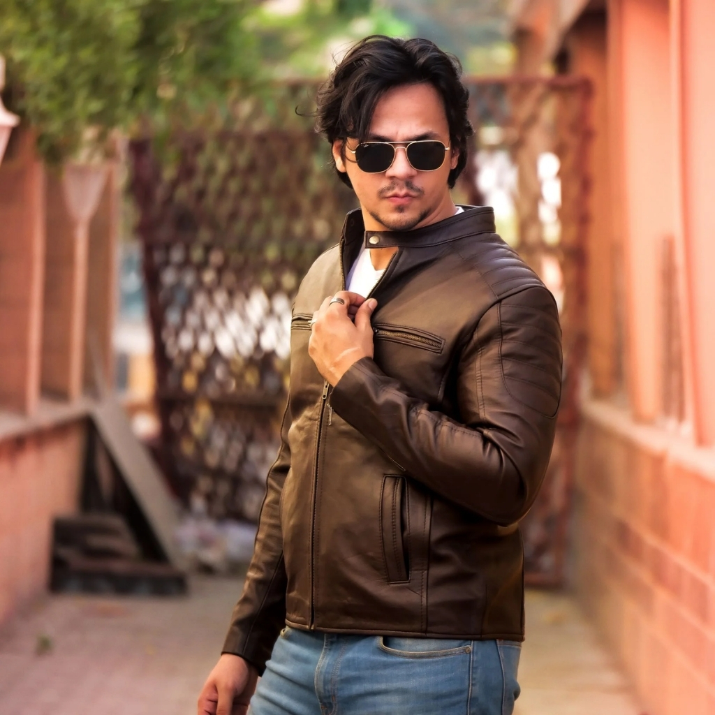 A man wearing a brown leather jacket, blue jeans, and sunglasses stands outdoors, adjusting his jacket.