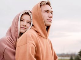A young couple wearing hoodies outdoors. The woman with light brown hair wearing a mauve hoodie leans her head on the shoulder of the man, who has short blond hair and a brown hoodie. They stand in a grassy, tree-dotted field under a cloudy sky.