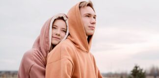 Streetwear Hoodie: Trendy Urban Style for Every Wardrobe A young couple wearing hoodies outdoors. The woman with light brown hair wearing a mauve hoodie leans her head on the shoulder of the man, who has short blond hair and a brown hoodie. They stand in a grassy, tree-dotted field under a cloudy sky.