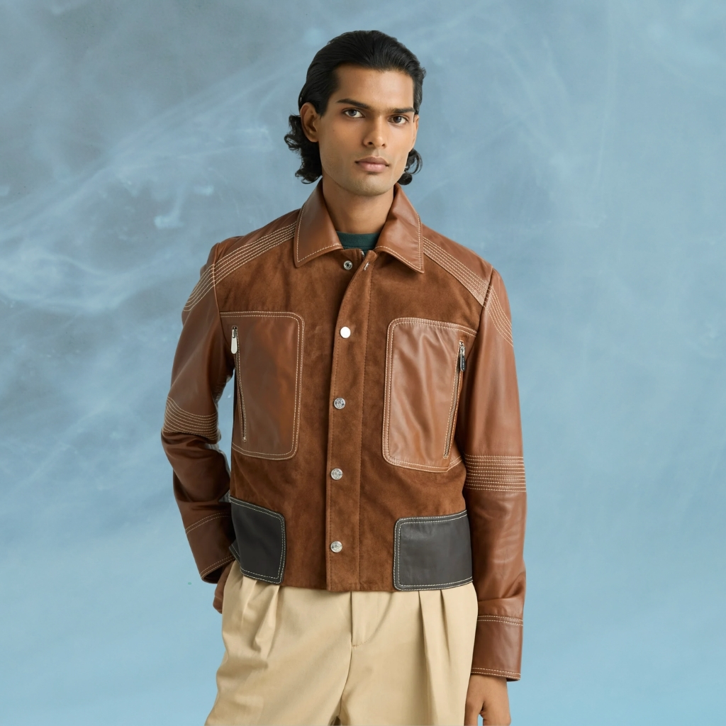 A slender man with dark hair stands against a soft, smoky blue backdrop, wearing a brown paneled jacket made of suede and smooth leather with large zip pockets and dark leather panels at the hem, paired with beige pleated trousers.