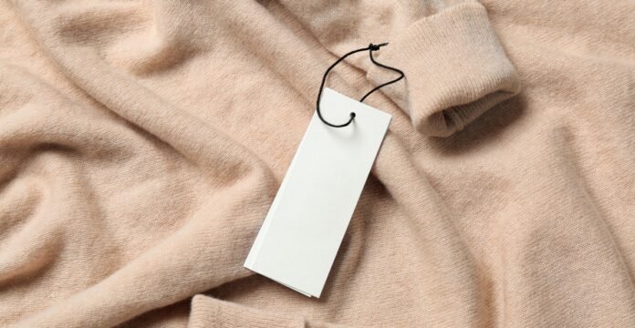 Sustainable Grade B Cashmere Close-up of a light peach cashmere sweater with a blank white tag hanging from a black string, casually folded to show a cuff in the upper right corner.