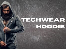 A bearded man in a gray zip-up hoodie stands against a textured gray background, partially zipping up the hoodie. Text on the right reads “TECHWEAR ZIP HOODIE.”