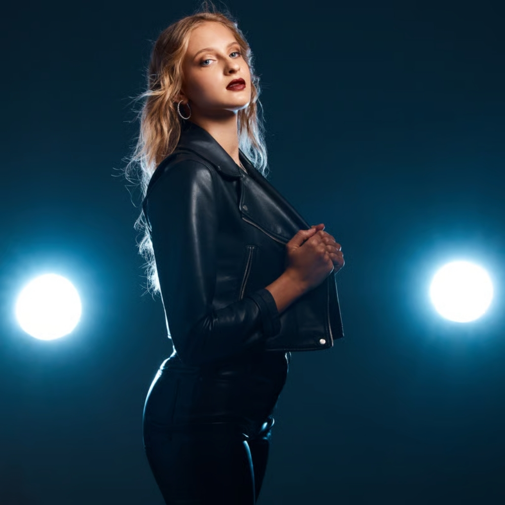 A young woman with long blonde hair stands in front of a dark blue background, wearing a black leather jacket and black pants. Two bright circular lights are visible on either side of her, and she looks to the left with a serious expression.