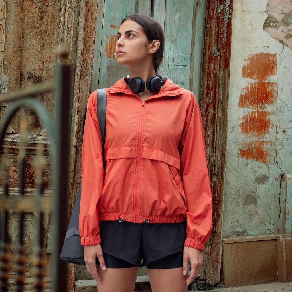 A young woman stands outdoors in an urban setting, wearing a bright red CityTrek jacket, black athletic shorts, and a gray bag over her shoulder. She has a pair of black headphones around her neck and gazes upward. The weathered door and faded wall behind her show wear and age.