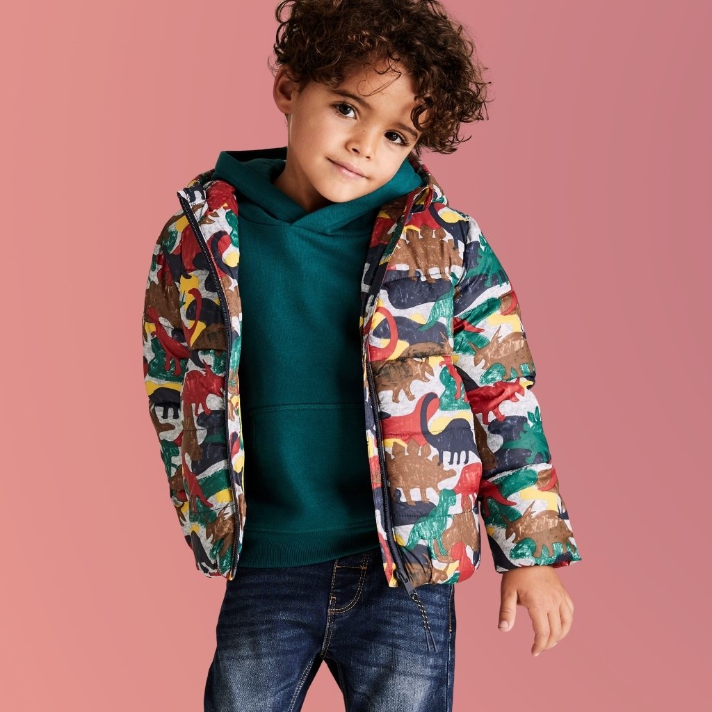 A young boy with curly brown hair stands against a pink background, wearing a green hoodie underneath a colorful dinosaur-patterned jacket, and dark blue jeans. The jacket is unzipped, revealing the hoodie, and the boy has a gentle, off-center gaze.Dino Dash Jacket for Kids