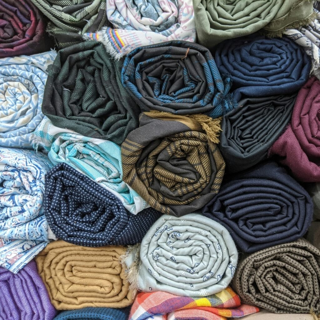 The Essential Pullover Plain Hoodie: Style GuideĀ 4 A dense arrangement of rolled fabrics in assorted colors and patterns, showing blue, brown, green, purple, and red hues with plaids, paisleys, solids, and textured weaves; some rolls have fringe. Pullover plain hoodie