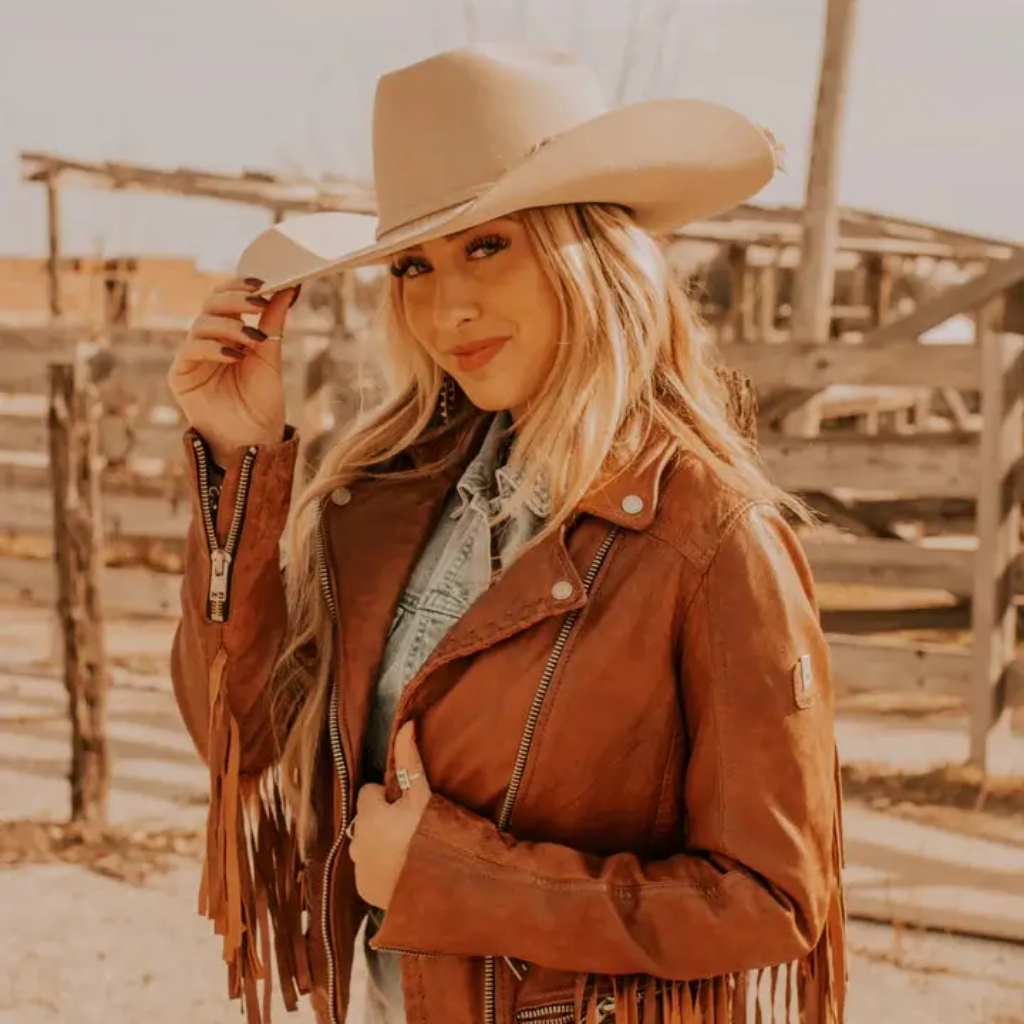 Stylish Western Leather Jacket Inspired by Classic Cowboy Wear 4 A smiling woman wearing a tan cowboy hat and a brown leather fringed jacket stands beside a wooden fence. She holds the hat brim with one hand, and sunlight gives a warm, golden glow to the scene.Stylish Western Leather Jacket