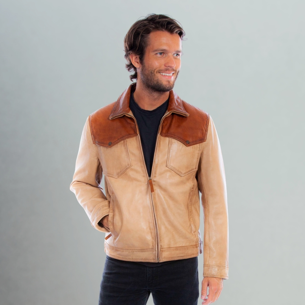Two-Tone Leather Jacket for Bold Modern Everyday Street Style 5 A light-skinned man with brown hair and a slight smile stands against a gray background, wearing a two-tone brown leather jacket over a black T-shirt and dark jeans. His left hand is in his pocket.
