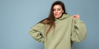 Typography Hoodie: Bold Style Meets Street Fashion A young woman with long brown hair wearing an oversized light green hoodie, standing against a solid blue background; she has a slight smirk and looks at the viewer.