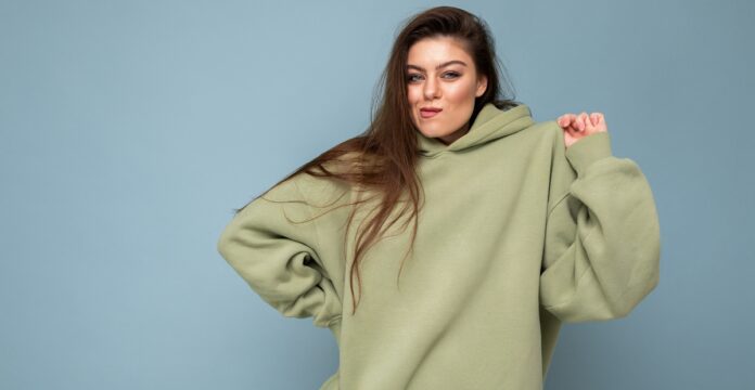 A young woman with long brown hair wearing an oversized light green hoodie, standing against a solid blue background; she has a slight smirk and looks at the viewer.
