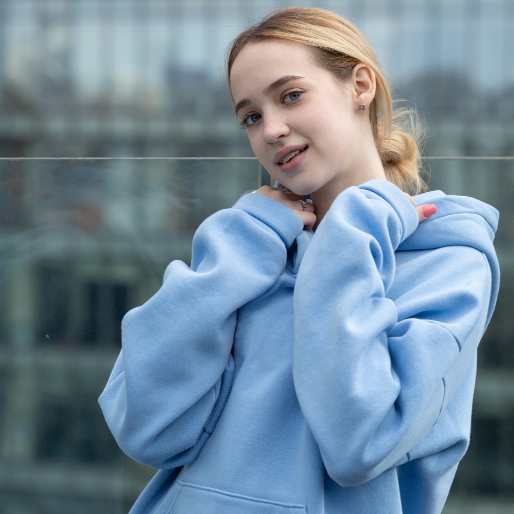 A young woman with blonde hair in a low bun wearing a light blue hoodie. She crosses her arms near her neck and smiles softly at the camera, with a blurred cityscape behind her.
typography hoodie
