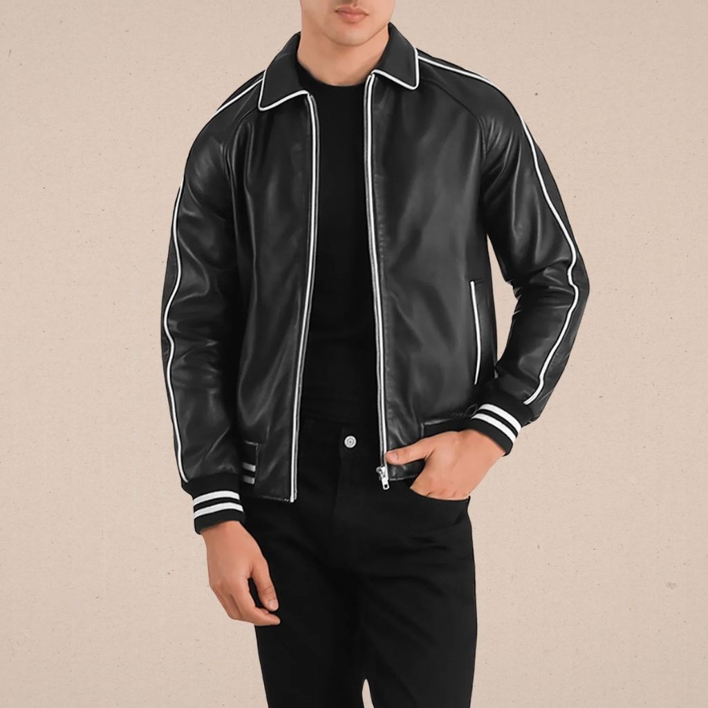 A man stands facing the camera wearing a black leather varsity jacket with white piping, cuffs, and waistband, zipped up over a black shirt, paired with black pants. The background is a plain light brown.